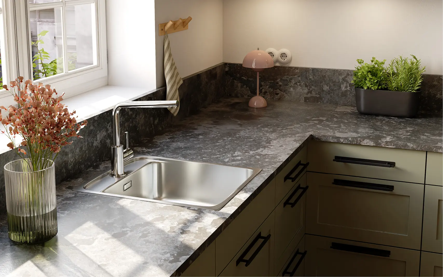 Detailansicht Küchenarbeitsplatte mit dunkler Steinoptik: Edelstahlspüle mit Chromarmatur unter Fenster, Glasvase mit Blumen, kleine rosa Tischlampe, Kräutertopf; salbeigrüne Rahmenfronten mit schwarzen Griffen.